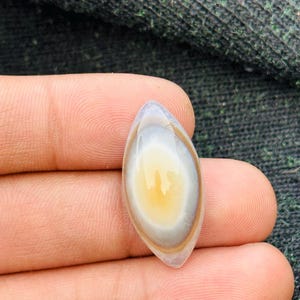 Könnte beinhalten: Ein polierter, oval geformter Edelstein mit einem gelben Kern, umgeben von konzentrischen Bändern in Weiß, Braun und Grau. Der Stein wird in einer Hand gehalten, wodurch seine glatte, reflektierende Oberfläche hervorgehoben wird. Das Design des Edelsteins ähnelt einem Auge.