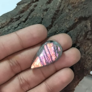 Puede incluir: Una gema pulida en forma de lágrima. La piedra muestra un vibrante juego de colores, con vetas rosas, moradas y naranjas. La gema se sostiene en una mano, mostrando su tamaño y forma.