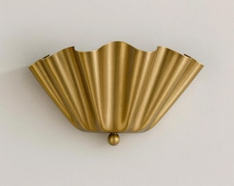May include: A gold-colored wall sconce with a scalloped, fan-like design. The sconce has a textured surface and a small, round detail at the bottom. The fixture is mounted on a white wall.