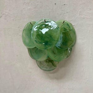 May include: A wall-mounted light fixture featuring a cluster of circular, translucent green glass discs with a marbled pattern. The discs are arranged in a layered, overlapping formation, creating a textured surface that diffuses light.