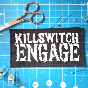 May include: A black rectangular patch with the words "KILLSWITCH ENGAGE" in white, surrounded by sewing tools. The tools include silver scissors, pins, safety pins, and clear clips, all arranged on a blue cutting mat.