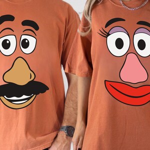 May include: Two orange t-shirts with cartoon faces printed on them. One shirt features a male character with a tan nose, black mustache, and a smiling mouth. The other shirt has a female character with a pink nose, red lips, and eyelashes.