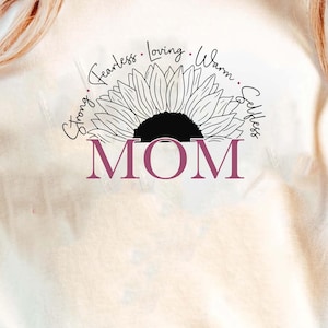 May include: Cream-colored t-shirt featuring a sunflower design with the word "MOM" in a burgundy font. The sunflower is outlined in black, with the words "Strong, Fearless, Loving, Warm, Selfless" arching above the flower.