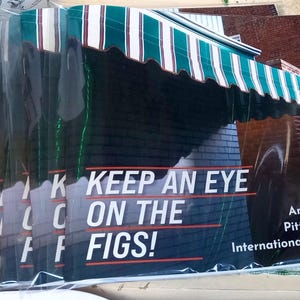 May include: A stack of promotional posters featuring a storefront with a green and white striped awning. The posters have the text "KEEP AN EYE ON THE FIGS!" and "An Ode to Pittsburgh's International Grocers."