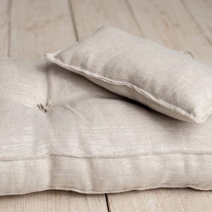 May include: A rectangular beige cushion with a tufted surface and visible stitching along the edges, paired with a smaller, smooth rectangular beige pillow resting on top. Both items feature a woven texture and a neutral, light tan colour palette.