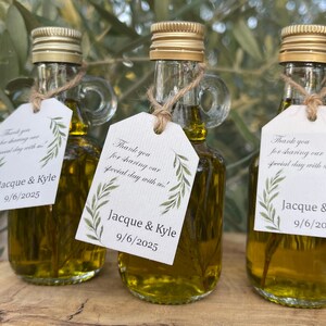 Könnte beinhalten: Drei kleine Glasflaschen gefüllt mit Olivenöl, jede mit einem goldenen Verschluss und einem Griff. Jede Flasche hat ein Etikett, das mit Bindfaden befestigt ist und "Danke, dass Sie unseren besonderen Tag mit uns geteilt haben!" und die Namen "Jacque & Kyle" mit dem Datum "9/6/2025" trägt.