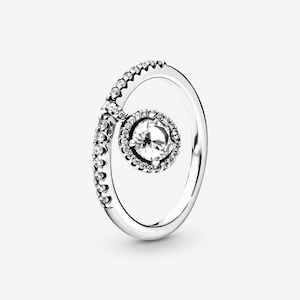 May include: A silver ring with a large, clear, round gemstone at the center, surrounded by smaller clear stones. The band curves around the main stone, with a section of the band also set with small, clear stones. The ring is set against a white background.