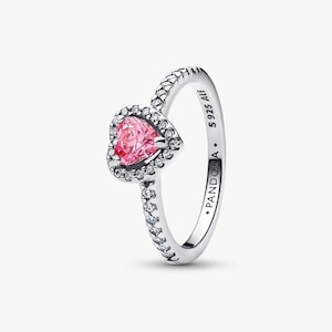 Puede incluir: Un anillo plateado con una piedra rosa en forma de corazón en el centro, rodeada de piedras claras más pequeñas. La banda está detallada con pequeñas cuentas en relieve. El anillo está marcado con "PANDORA" y "S925 ALE".