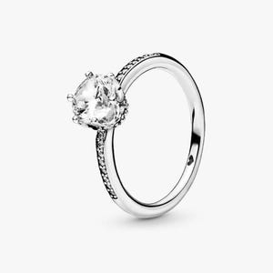 May include: A silver ring featuring a large, clear, faceted gemstone. The band is smooth, with a row of small, clear stones set along the top. The ring is set against a white background. The ring's design is elegant.
