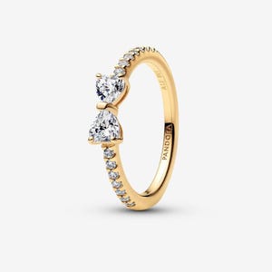 Puede incluir: Un anillo dorado con diseño de lazo. El anillo presenta dos piedras claras en forma de corazón y una fila de piedras más pequeñas. La banda es lisa y pulida, con el nombre de la marca "PANDORA" grabado en el lateral.