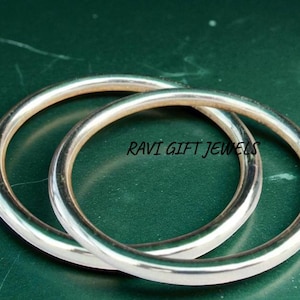 May include: Two silver bangles, circular in shape, are displayed against a dark green background. The bangles are smooth and shiny, reflecting light. The text "RAVI GIFT JEWELS" is visible on the surface. These are likely jewelry items.