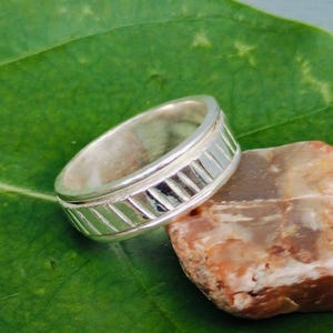 Peut inclure: Une bague en argent avec un motif géométrique repose sur une petite pierre brun-rouge. La bague est posée sur une grande feuille verte vibrante, créant un contraste saisissant. Le motif de la bague présente une série de lignes parallèles.