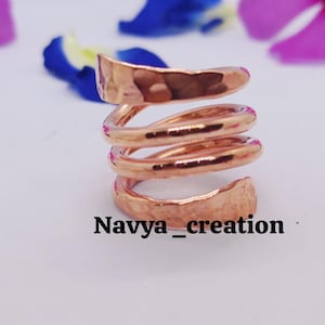 May include: A copper-colored spiral ring with a hammered texture. The ring wraps around the finger multiple times, creating a unique and eye-catching design. The text "Navya_creation" is at the bottom of the image. The background includes flower petals.