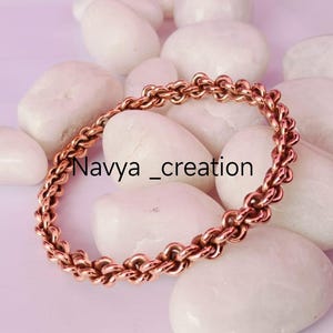 May include: A copper-colored chain bracelet with a linked design. The bracelet is displayed on a bed of smooth, white stones against a soft purple background. The text "Navya_creation" is visible.