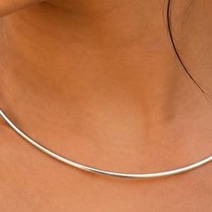 May include: A smooth, circular silver-toned metal choker necklace that rests around the neck. The piece features a simple, continuous band design with a polished, reflective finish, maintaining a uniform thickness throughout its entire circumference.