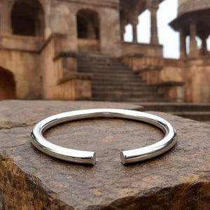 May include: A shiny, silver-colored open cuff bracelet. The bracelet is a thick, rounded band with a smooth, polished surface. The ends of the bracelet are open, creating a gap. The bracelet is resting on a textured, brown stone surface.