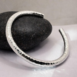May include: A silver-colored cuff bracelet with a hammered texture. The open-ended bracelet is curved and rests on a dark gray stone. The bracelet's surface reflects light, highlighting its textured design. This piece is a type of jewelry.