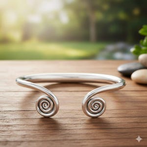 May include: A silver-colored cuff bracelet with a unique design. The bracelet features a curved band that ends in spiral designs on each end. The bracelet is displayed on a wooden surface with a blurred green background.