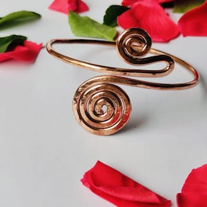May include: A copper-colored arm cuff with spiral designs. The cuff features a large spiral at the center and a smaller spiral at the top. Red rose petals and green leaves are scattered around the cuff on a white surface.