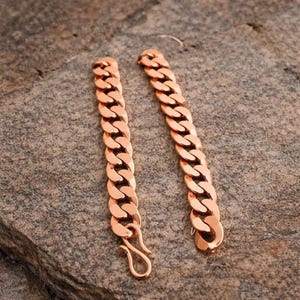 May include: Two copper-colored chain bracelets with a curb link design. One bracelet has a hook clasp, and the other has a loop. The bracelets are displayed on a gray stone surface.
