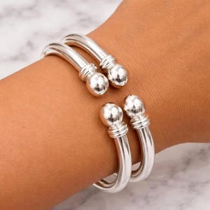 Pode incluir: Duas pulseiras de punho abertas prateadas. Cada pulseira tem uma extremidade arredondada e bulbosa e uma faixa decorativa perto da extremidade. As pulseiras são usadas em um pulso, mostrando seu design e brilho metálico.