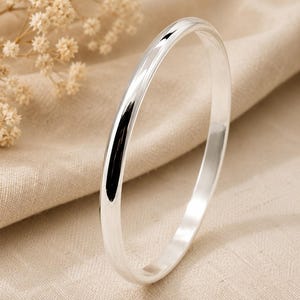 Peut inclure: Un bracelet jonc en argent poli avec une surface lisse et arrondie. L'anneau circulaire présente une finition métallique réfléchissante qui capte la lumière, créant un aspect propre et simple. Le bracelet est un anneau solide et continu.