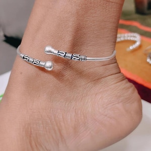 May include: A silver-toned open-ended anklet featuring a smooth, rounded ball at each terminal end. The band is decorated with etched, dark-colored geometric patterns and horizontal line detailing near the ends, creating a textured appearance.