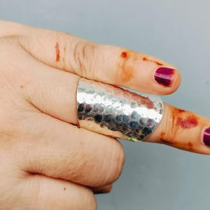 Può includere: Un anello largo e argentato con una texture martellata. L'anello è indossato su un dito e le unghie sono dipinte di un colore viola scuro. Lo sfondo è di colore grigio chiaro.