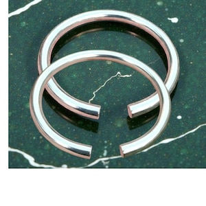 May include: Two silver-colored, open-hoop earrings. The earrings are circular with a small gap at the bottom. The earrings are displayed on a dark green, speckled surface. The earrings are made of a shiny metal.