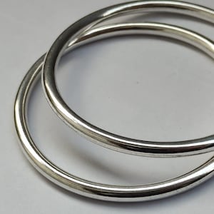 Puede incluir: Dos brazaletes circulares entrelazados con un acabado plateado liso y pulido. Los brazaletes son delgados, de sección transversal redondeada y presentan una superficie metálica reflectante que capta la luz, creando un diseño de bucle.