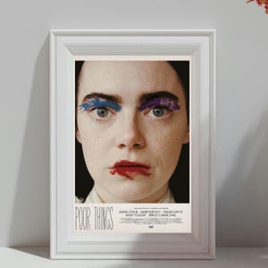 May include: Framed movie poster for "Poor Things" featuring Emma Stone. The poster shows a close-up of a woman's face with blue and purple eye makeup and red lipstick. The title and cast names are at the bottom.