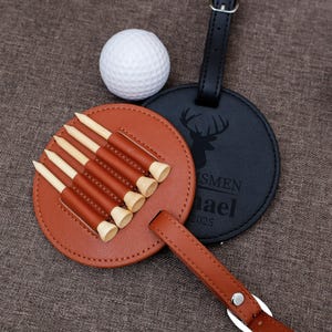 Puede incluir: Un juego de regalo temático de golf con una pelota de golf blanca, un soporte para tee de cuero marrón con tees de madera y una etiqueta de equipaje de cuero negro con una silueta de ciervo. El soporte para tee tiene un asa y contiene seis tees.