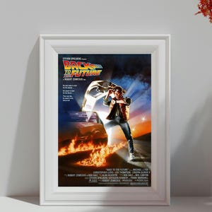 Puede incluir: Póster enmarcado de la película "Regreso al Futuro" con Michael J. Fox. El póster muestra un coche DeLorean envuelto en llamas con un hombre con chaleco y vaqueros. El título está en una fuente estilizada con un diseño futurista.
