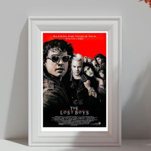 Puede incluir: Póster enmarcado de la película "The Lost Boys" con fondo rojo. El póster presenta a varios actores, incluido uno con gafas de sol. El texto en la parte superior dice: "Sleep all day. Party all night. Never grow old. Never die. It's fun to be a vampire."