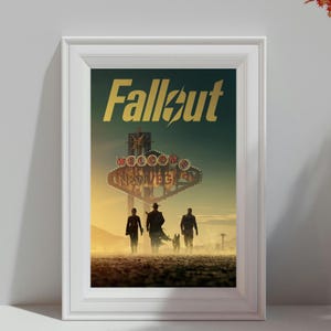 May include: A framed poster featuring the Fallout logo in gold text above a desert landscape. The scene shows a Welcome to New Vegas sign and three silhouettes walking across the dusty ground under a hazy, golden-toned sky at sunset.