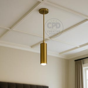 Puede incluir: Una lámpara colgante cilíndrica de color dorado cuelga de un techo blanco. La luz tiene un diseño simple y moderno, con una base redonda y una pantalla cilíndrica. El accesorio está suspendido por una fina varilla dorada. La habitación tiene una paleta de colores neutros.