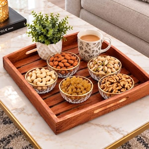 Könnte beinhalten: Ein quadratisches Holztablett mit Lattenseiten und Griffaussparungen, das fünf kleine, klare Glasschalen mit Goldrand enthält, gefüllt mit Mandeln, Cashews, Rosinen, Pistazien und Walnüssen, sowie eine weiße Keramiktasse und eine kleine Topfpflanze.