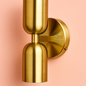 Könnte beinhalten: Eine goldfarbene Wandleuchte mit zylindrischem Design. Die Leuchte hat einen abgerundeten Sockel und einen zylindrischen oberen Teil, verbunden durch einen kurzen, zylindrischen Abschnitt. Die Leuchte ist auf einer kreisförmigen Platte montiert. Der Hintergrund ist hellrosa.