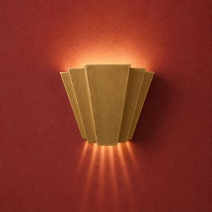 Könnte beinhalten: Eine Wandleuchte mit fächerförmigem, gestuftem Design aus gebürstetem Goldmetall. Die Leuchte strahlt an den oberen und unteren Kanten ein warmes, leuchtendes Licht aus und wirft einen sanften Schein auf die strukturierte, tiefrote Wand.