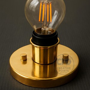 Könnte beinhalten: Eine Vintage-Glühbirne mit einem transparenten Glaskolben und warmen orangefarbenen Filamenten. Sie ist auf einem goldfarbenen Metallsockel mit schwarzem Akzent und einem kleinen Knopf montiert. Der Sockel trägt den Text "CREATIVE POINTS DESIGN".