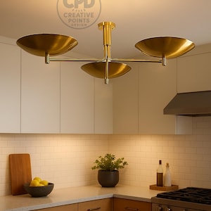 K&ouml;nnte beinhalten: Eine goldfarbene Deckenleuchte mit drei flachen, schalenf&ouml;rmigen Lampenschirmen, die von einem zentralen vertikalen Stab ausgehen. Die Metallstruktur hat eine polierte Oberfl&auml;che und das Design enth&auml;lt ein kreisf&ouml;rmiges Logo mit dem Text CPD CREATIVE POINTS DESIGN.