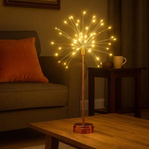 Op de afbeelding: Een decoratieve tafellamp met een sterrenuitbarsting ontwerp. De lamp heeft een bronzen basis en steel, met talrijke kleine, warmwitte LED-lampjes die naar buiten stralen. De lamp staat op een houten tafel in een woonkamer.