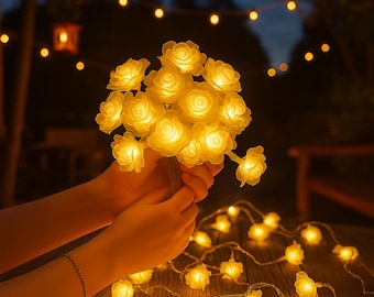 May include: A string of illuminated rose-shaped lights, held in a person's hands, with a second string of lights on a wooden surface. The lights are a warm yellow color, creating a soft glow against the dark background. The overall theme is decorative lighting.