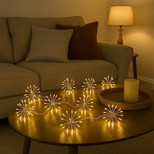 Op de afbeelding: Een reeks warmwitte sterrenburst-lampjes op een houten salontafel. De lampjes zijn in een rij gerangschikt, waarbij elke ster een zachte gloed uitstraalt. De tafel staat in een woonkamer met een bank en een lamp.