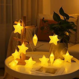 Op de afbeelding: Een reeks warmgele stervormige feeërieke lichtjes, waarvan sommige op een witte tafel liggen. De lichtjes zijn gerangschikt met een houten decoratiestuk en een plant in een pot op de achtergrond.