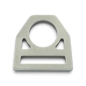 Puede incluir: Una pieza de herraje metálico de color plateado, plana y con forma trapezoidal de esquinas redondeadas. Incluye una gran abertura circular en la parte superior, dos pequeños recortes triangulares en los laterales y una ranura rectangular ancha en la base.