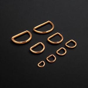 May include: An assortment of gold-coloured D-rings in different sizes, displayed on a black background. The metal D-rings have a smooth, polished finish. Sizes vary, suitable for crafts and hardware. The smallest ring is approximately 0.6 cm in diameter.