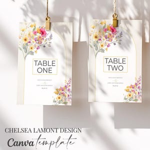 May include: Three hanging wedding table cards with floral designs. Each card is held by a gold clip and twine. The cards feature the words "TABLE ONE", "TABLE TWO", and "TABLE THREE" in a gold font. The bottom of the image has the text "CHELSEA LAMONT DESIGN" and "Canva template".