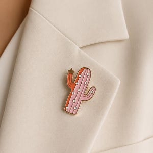 Puede incluir: Un broche de esmalte en tono dorado con forma de cactus, que presenta un diseño dividido con un lado en color naranja coral y el otro en rosa claro. El cactus tiene pequeños puntos blancos que representan espinas y una estrella dorada.