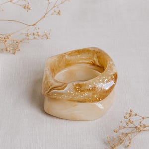 Puede incluir: Dos brazaletes de resina apilados con forma cuadrada redondeada. El brazalete superior es de color ámbar translúcido con remolinos blancos y purpurina incrustada, mientras que el inferior es de color crema opaco con un sutil veteado de color beige.